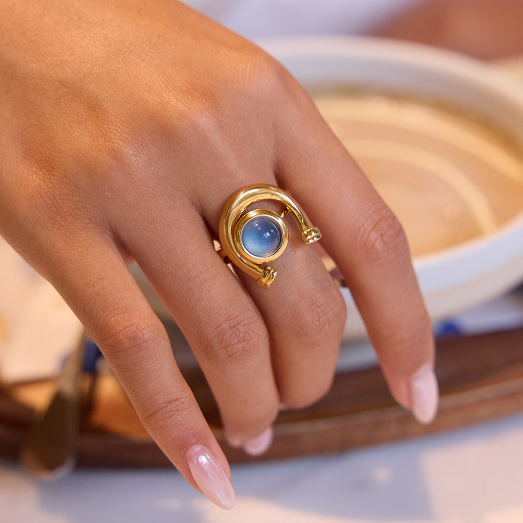Gold-tone stainless steel horseshoe ring with blue resin cabochon worn on a hand