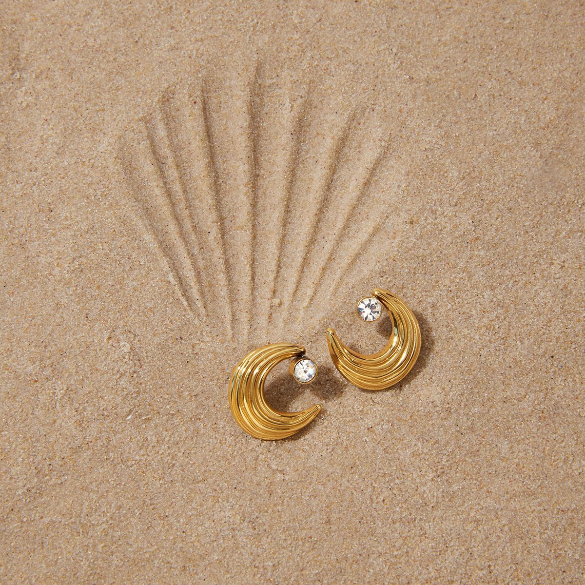 Gold-finish crescent studs with clear stones on a sand-colored surface