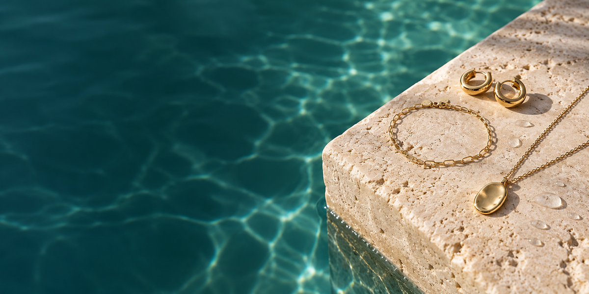Gold-tone jewelry beside a pool on a sunlit stone ledge